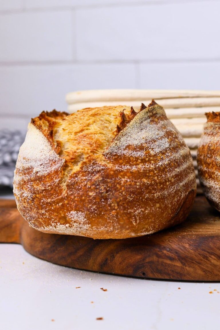 A loaf of golden sourdough bread with a double ear sitting on a wooden board. There is a banneton in the background of the photo.