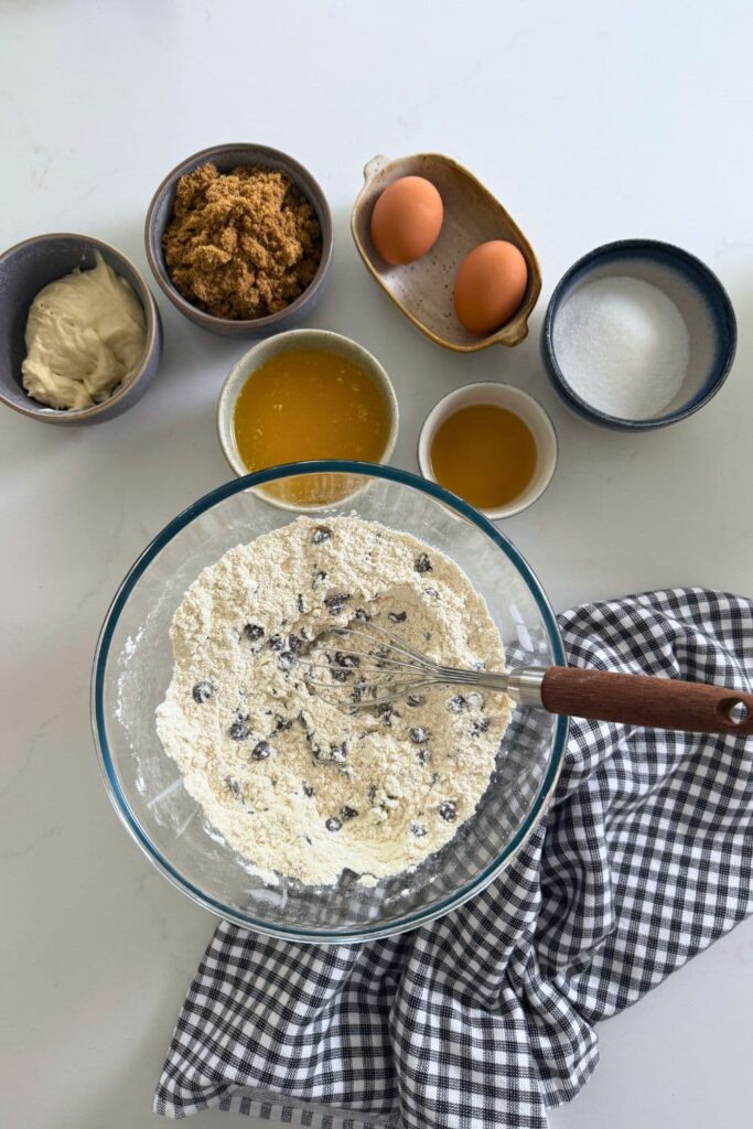 A glass bowl filled with flour and chocolate chips. There is a black and white dish towel wrapped around the glass bowl and the other wet ingredients in the photo with the bowl.