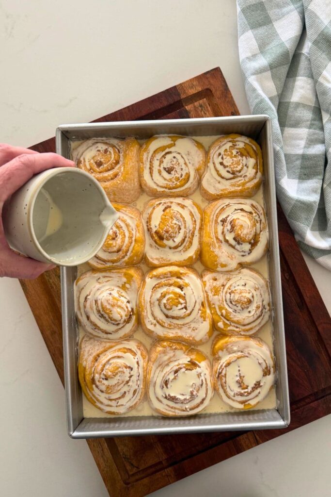 A jug of heavy cream being poured over a tray of sourdough cinnamon pumpkin rolls.