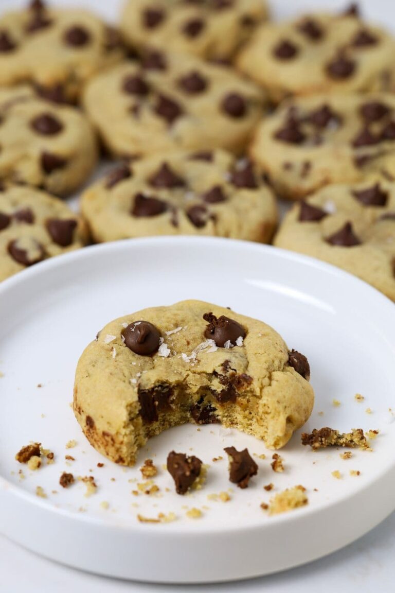 A sourdough chocolate chip cookie sitting on a white plate. There is a bite taken out of the cookie. There are lots of other cookies in the background of the white plate.
