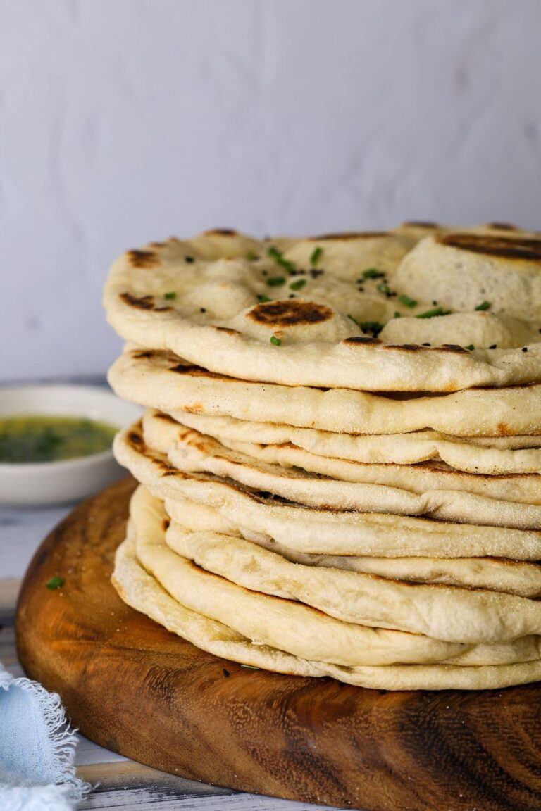 A stack of sourdough flatbreads on a round wooden board. There is a small dish of garlic butter in the background.