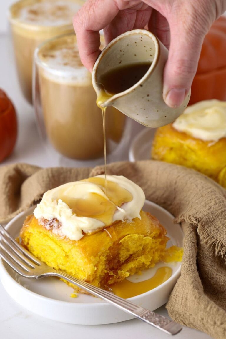 A sourdough pumpkin cinnamon roll smothered in vanilla cream cheese frosting being drizzled in maple syrup.