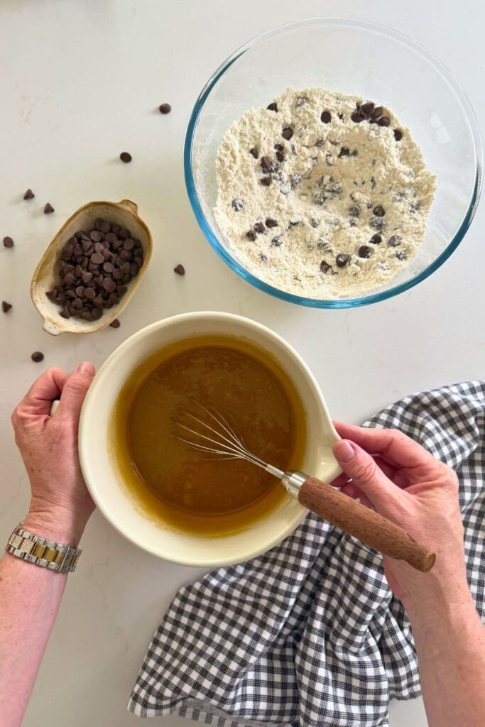 A pair of hands holding a cream batter jug filled the wet ingredients. There is a glass bowl filled with the dry ingredients sitting in the photo too.