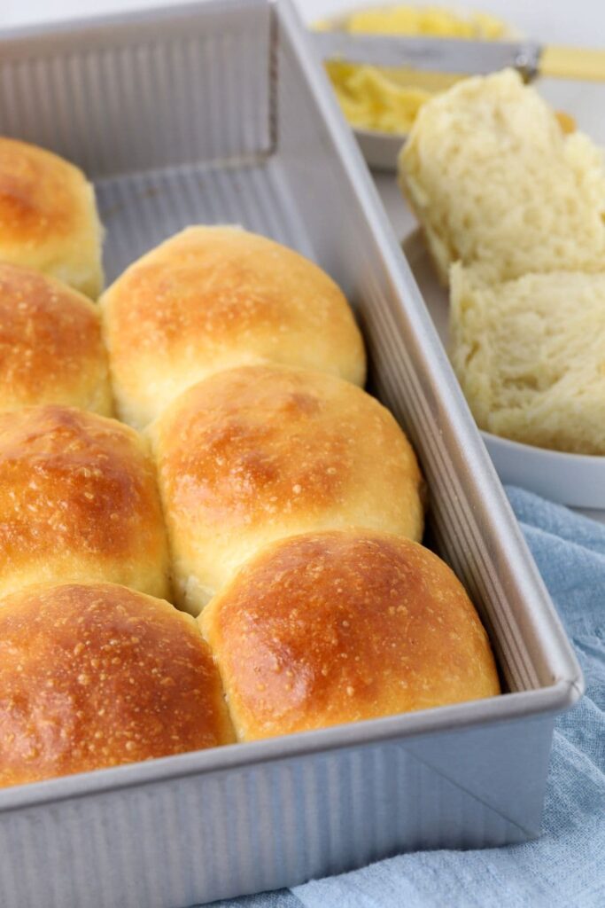 A baking pan of sourdough dinner rolls sitting on a counter top with a dinner roll cut open on a small plate in front of the tray.