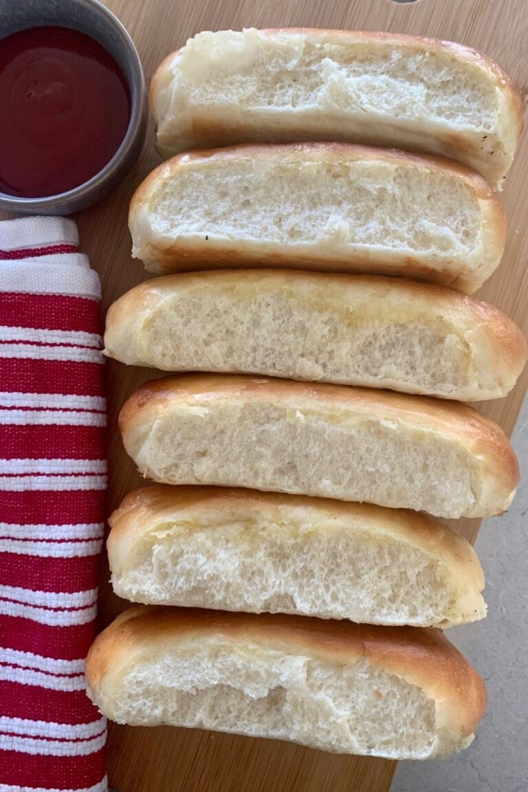 6 sourdough hot dog buns laying on their side so you can see their soft and squishy insides. There is a red striped dish towel next to them as well as a bowl of ketchup.