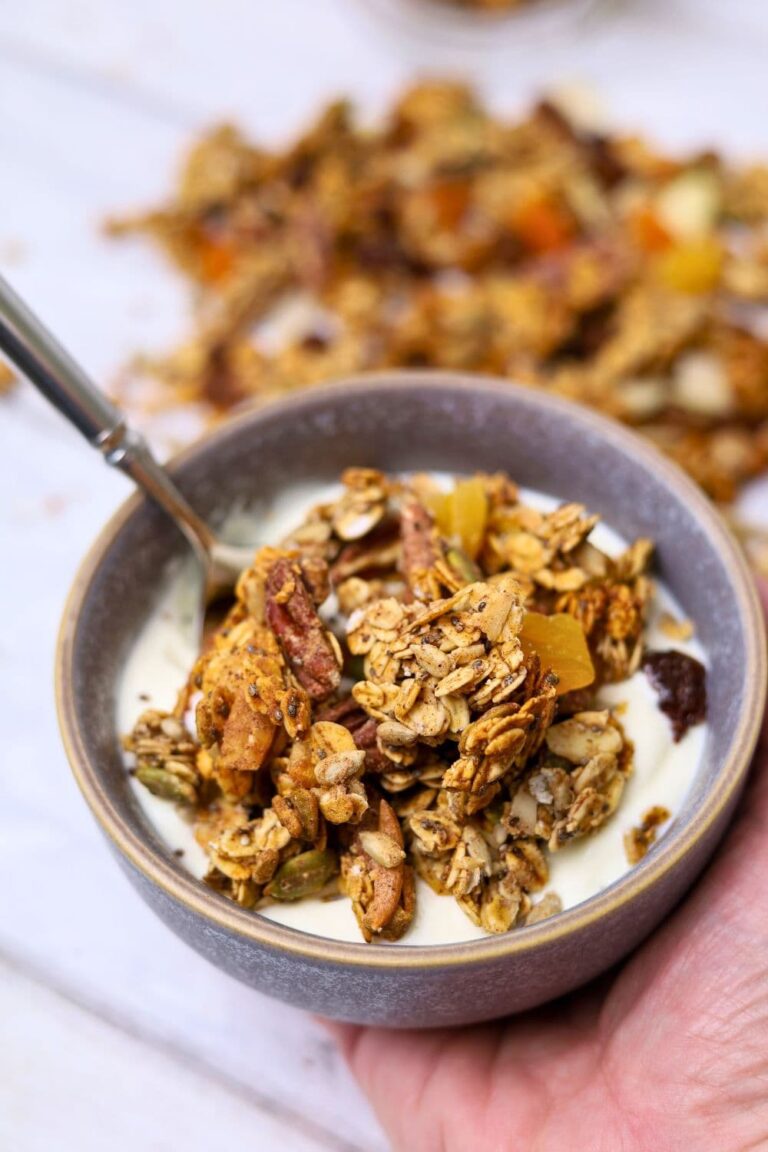 A hand holding a grey bowl of yogurt and sourdough granola. You can see more sourdough granola in the background of the photo.