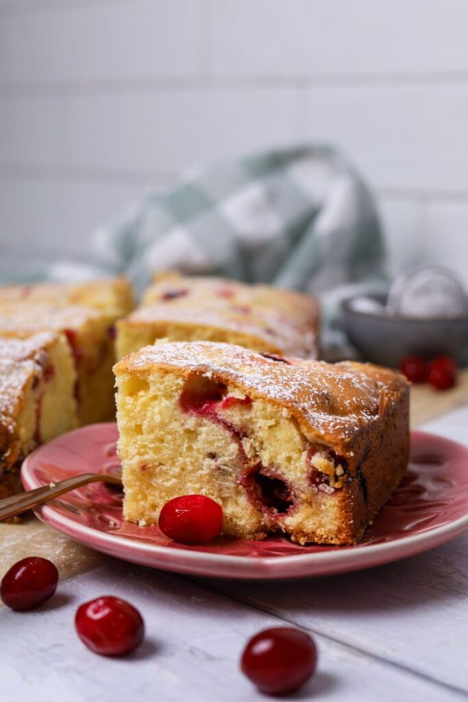 A delicate yellow sourdough cake studded with bright red cranberries. The cake is displayed on a small pink plate with a green and white checked dish towel in the background.