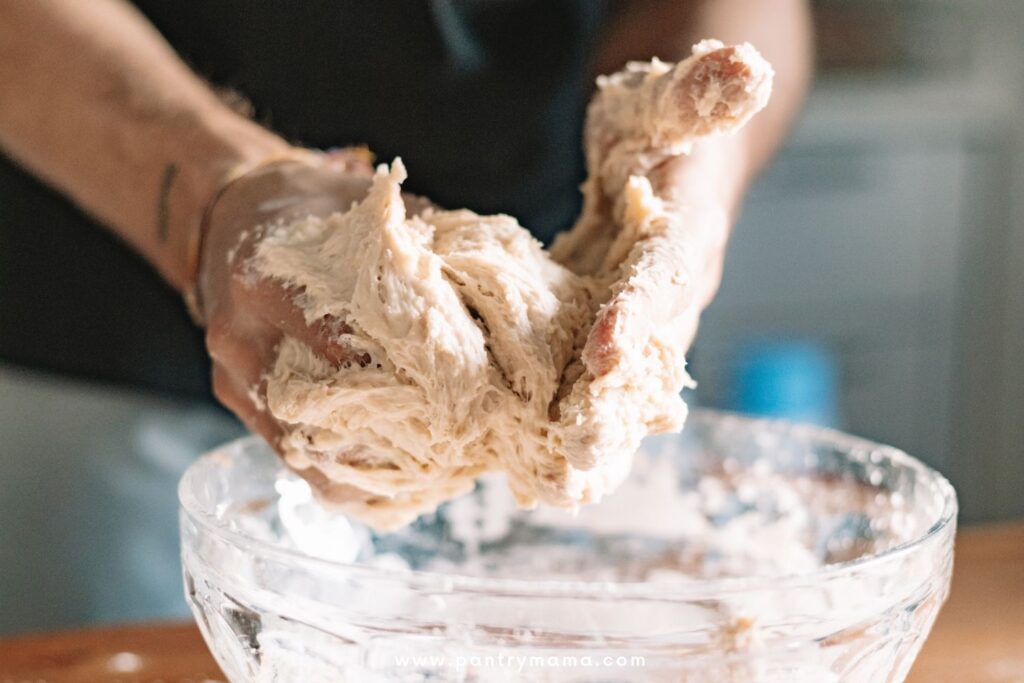 A pair of hands covered in wet, sticky sourdough hovering over a glass bowl.