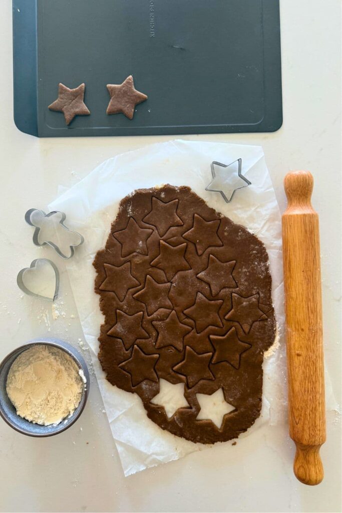 Sourdough gingerbread dough that has been rolled out onto a piece of parchment paper. 2 star shapes have been cut out of the dough and there is a rolling pin next to to the dough.