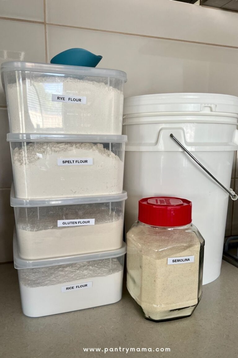 4 plastic containers with labelled with different types of flour stacked on top of one another. Next to them is a food safe bucket full of flour and a glass jar with a red lid containing semolina flour.
