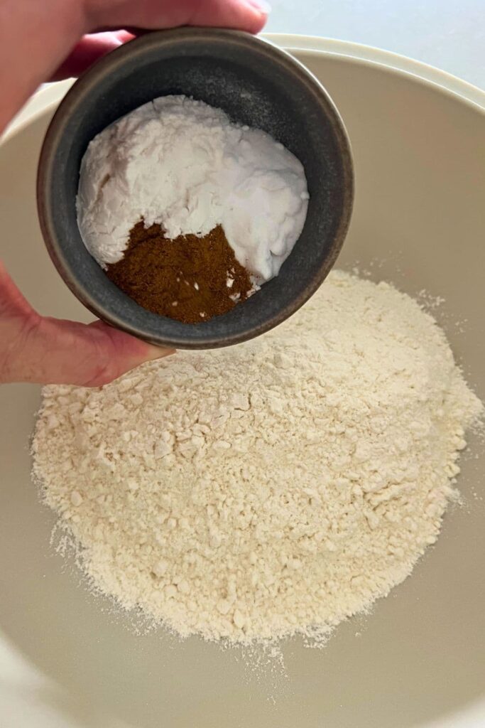 A small bowl containing cinnamon, cream of tartar, baking soda and salt about to be tipped into a big bowl of flour.