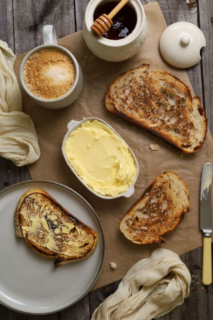 A flat lay with a small dish of homemade butter at the centre. The dish of butter is surrounded by 3 slices of toasted sourdough bread, a cup of coffee and a pot of honey.