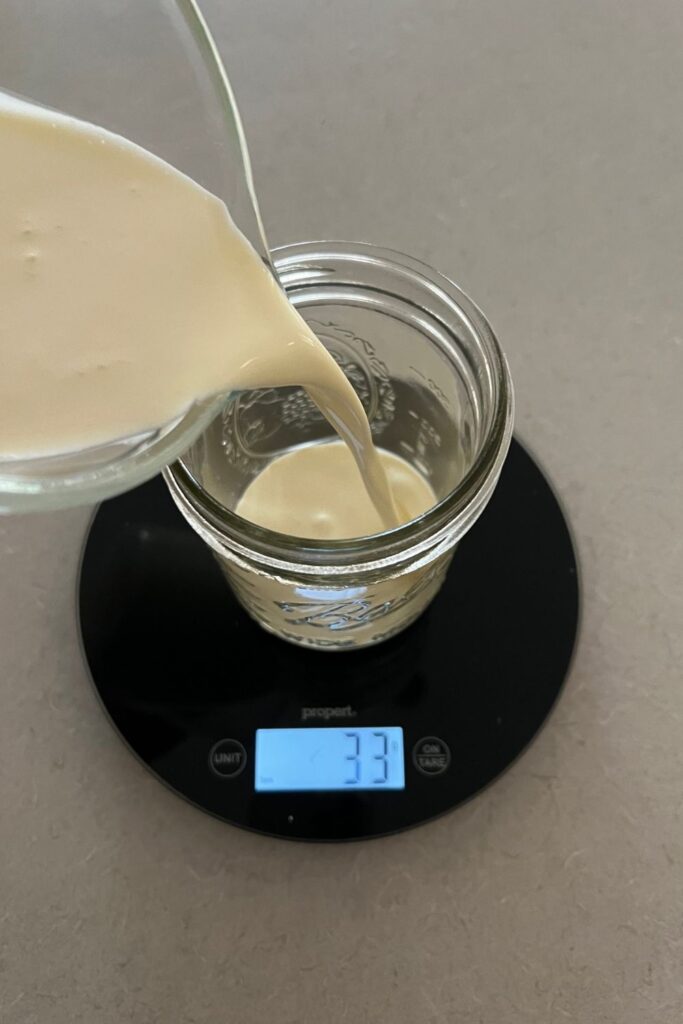 A mason jar sitting on a round black kitchen scale. Heavy whipping cream is being poured into the mason jar.