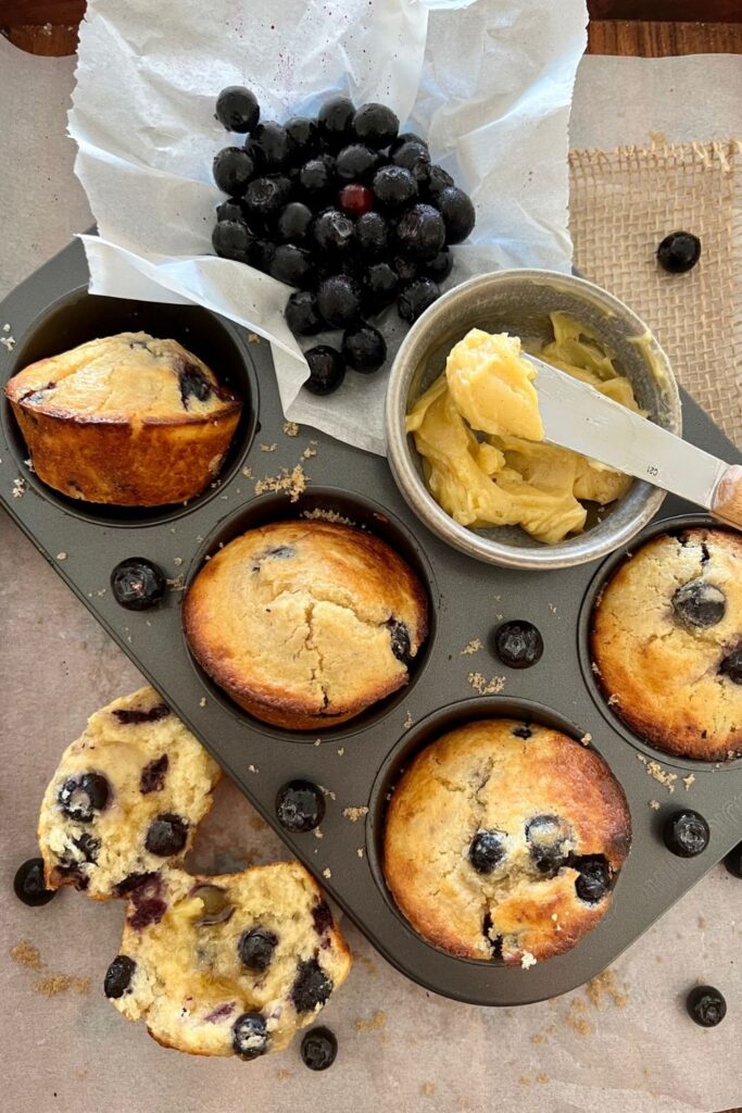 A 6 hole muffin tray containing sourdough blueberry muffins that have been baked. There is a muffin sitting to the left that has been broken open so you can see the inside of the muffin. There is also a pile of blueberries in the top right of the muffin pan. There is a dish of butter and a butter knife in the photo as well.