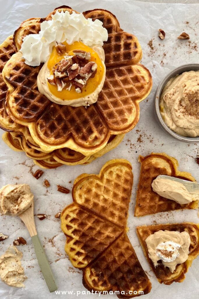 Sourdough pumpkin waffles stacked on a piece of parchment paper with some waffle pieces being topped with pumpkin spice cream cheese.