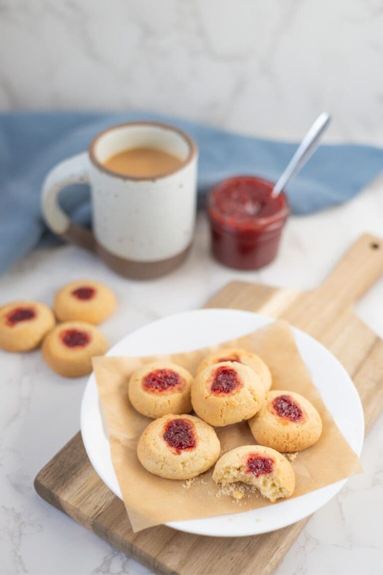 A white plate filled with sourdough discard thumbprint cookies. There is a cup of coffee and jar of strawberry jam in the background of the photo.