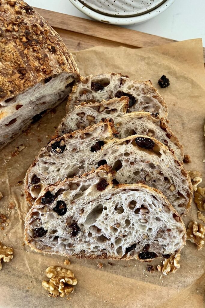 A loaf of walnut raisin sourdough bread that has been sliced and is sitting on a piece of parchment paper.