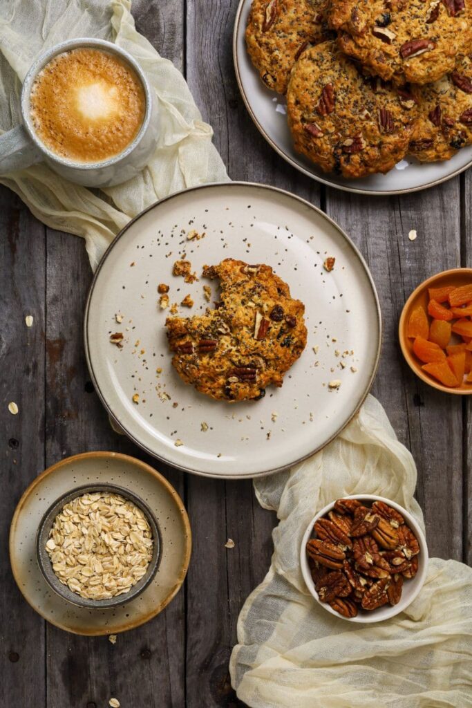 Flat lay of a rustic breakfast scene featuring a partially eaten sourdough breakfast cookie on a beige plate, surrounded by a cup of coffee, a bowl of dried apricots, a bowl of pecans, and a bowl of rolled oats on a dark wooden surface. A plate of more cookies and a soft cream-colored cloth add texture and warmth to the setup.