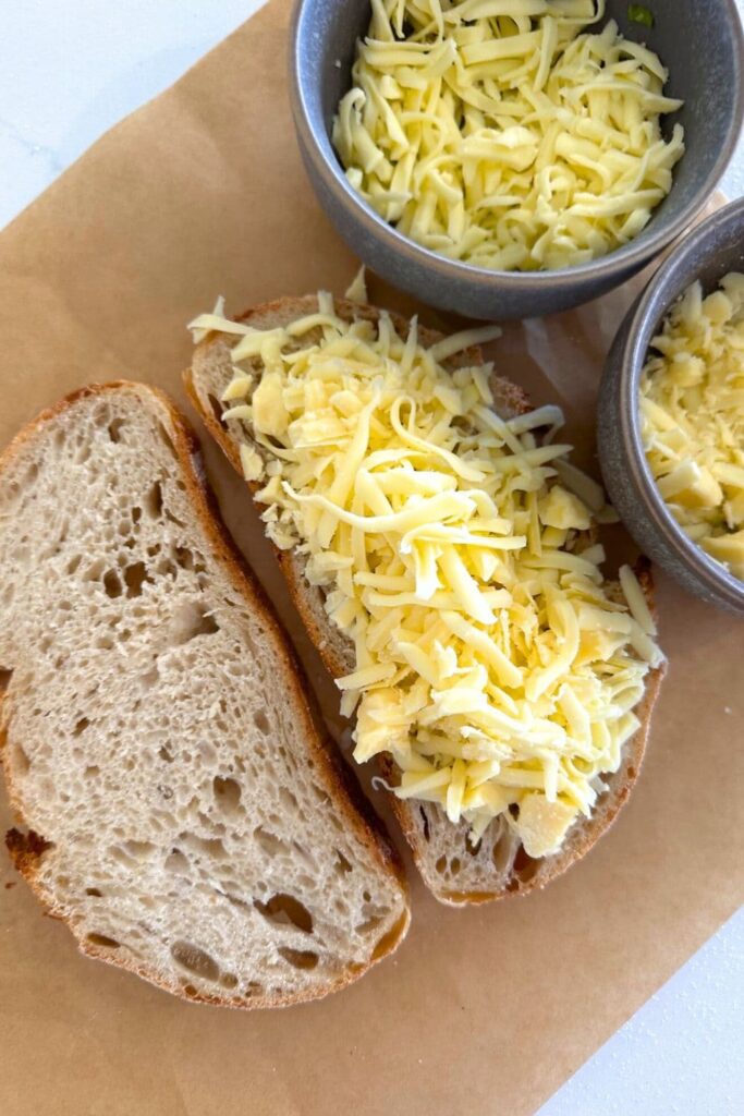 A photo showing two slices of sourdough bread. One slice is piled with shredded cheese. There are also two small bowls of shredded cheese to the right of the photo.