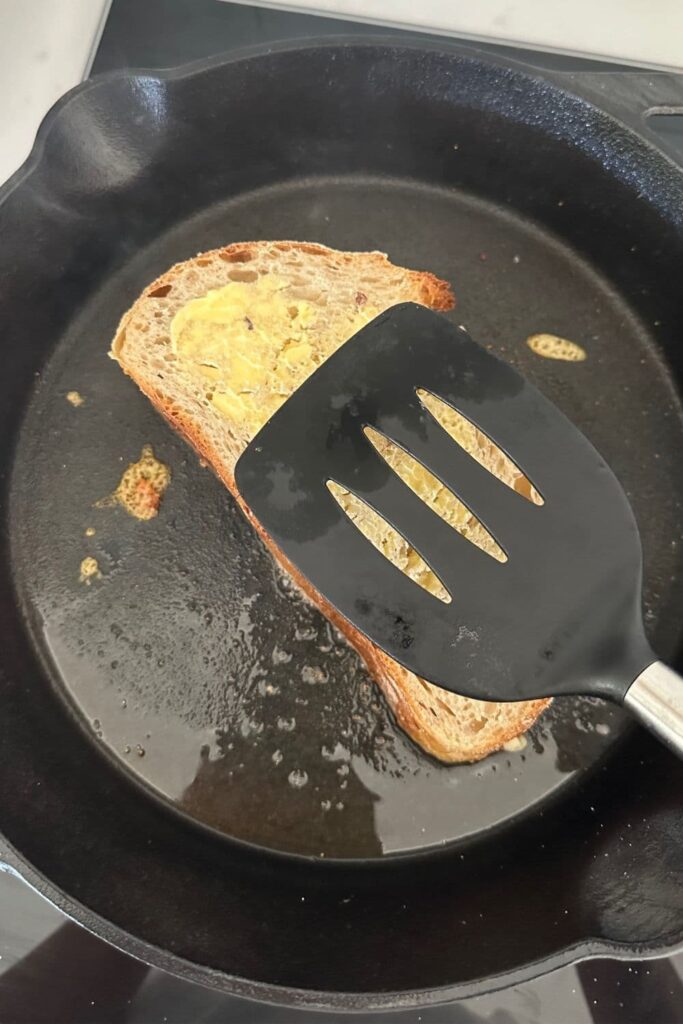 A sourdough grilled cheese being cooked in a cast iron skillet. There is a black spatula holding down the sandwich in teh pan.