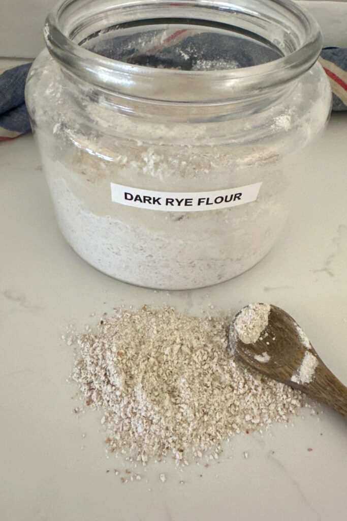 A large glass jar labelled "dark rye flour" sitting in front of a pile of dark rye flour that has been spilled on the counter top with a wooden spoon.