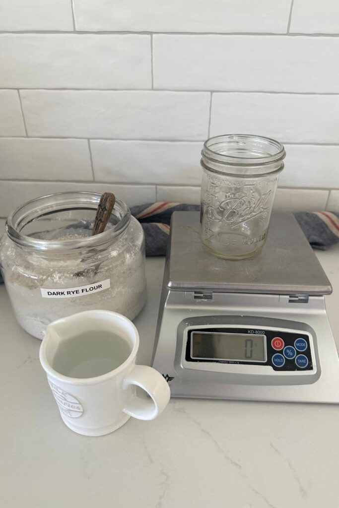 A digital kitchen scale sitting on a white counter top. There is an empty Ball jar sitting on top of the scale. To the left of the scale is a jar of dark rye flour and a small white jug of water.