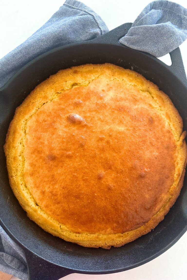 A cast-iron skillet containing sourdough cornbread that has just come out of the oven. It is golden brown and surrounded by a blue dish towel.