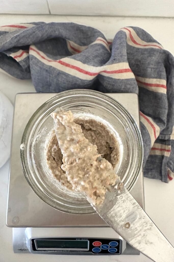A top down photo of a 16oz ball jar sitting on top of a kitchen scale. The ball jar contains a rye sourdough starter that has just been fed. Some of the rye starter is sitting on a knife that is sitting on top of the jar.