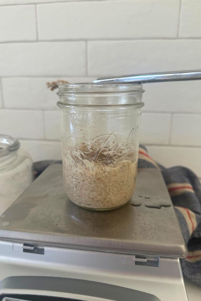 A jar of rye sourdough starter sitting on top of a kitchen scale. There is a knife sitting on top of the jar.