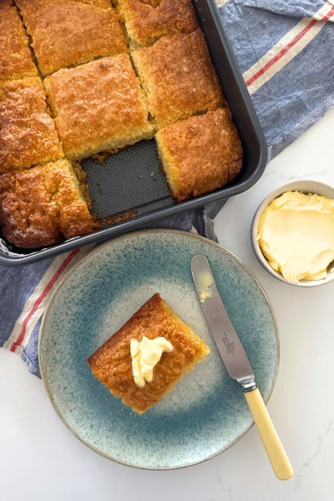 A square baking pan containing 8 sourdough butter swim biscuits. The 9th sourdough butter swim biscuit is sitting on a blue plate and topped with butter.
