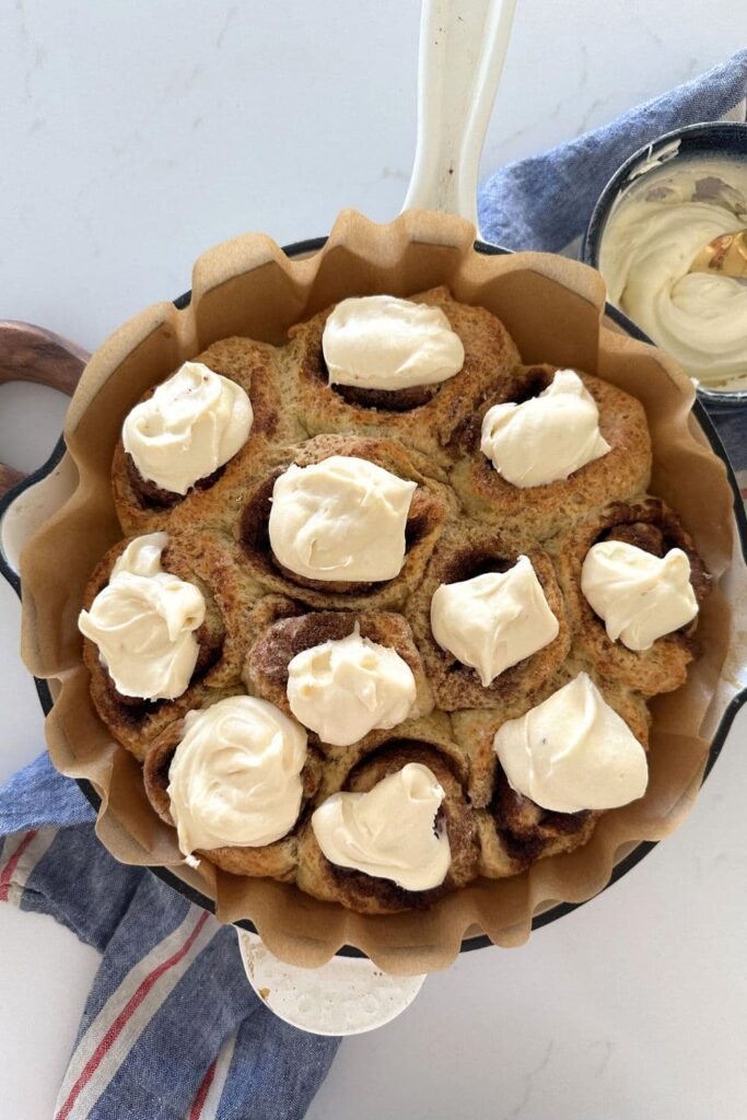 A cream enamel cast iron skillet filled with no wait sourdough cinnamon rolls topped with vanilla cream cheese frosting.