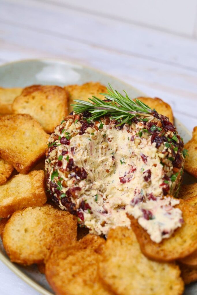 A cranberry pecan cream cheese ball served with sourdough crostini. The front of the ball has been dug into so you can see the delicious flavored cream cheese inside.