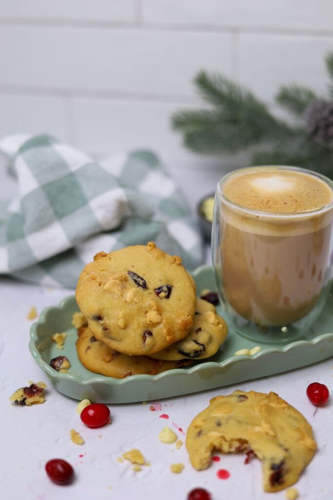 A green tray laid with a pile of sourdough white chocolate cranberry cookies. There is also a cup of coffee on the tray. In the foreground, there is a cookie with a bite taken out of it.