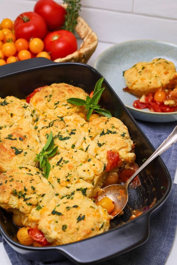 A black baking dish with sourdough tomato cobbler topped with fresh garlic and herb butter. There is a basket of red and yellow tomatoes in the background.