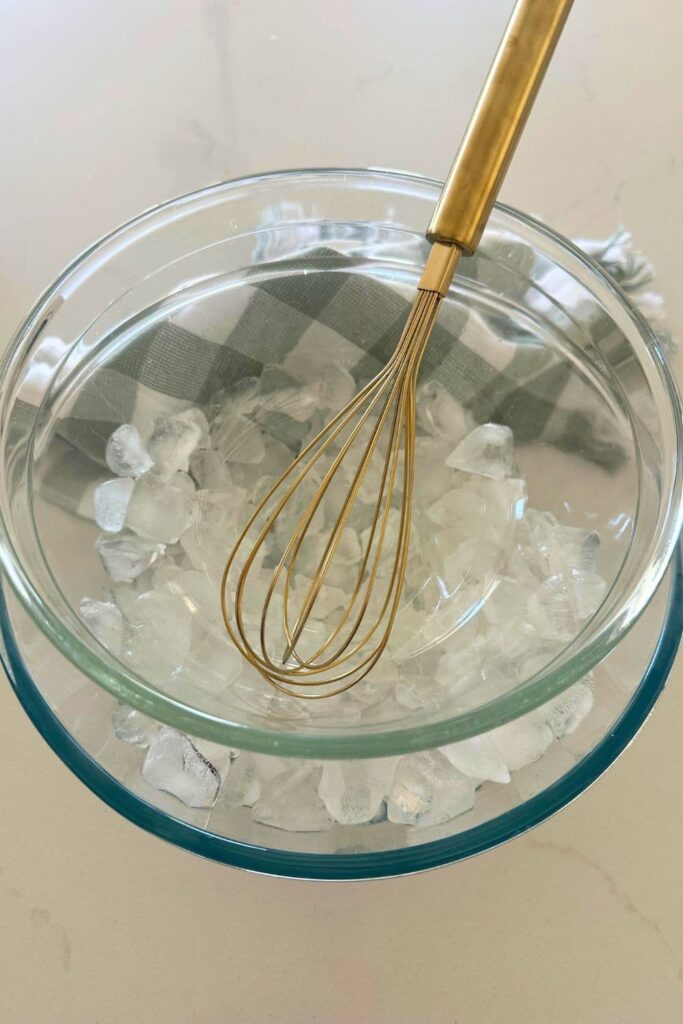 Two glass bowls nestled together. The bottom bowl is larger and contains ice. There is a gold whisk sitting in the top bowl and a green and white checked dish towel in the background of the image.