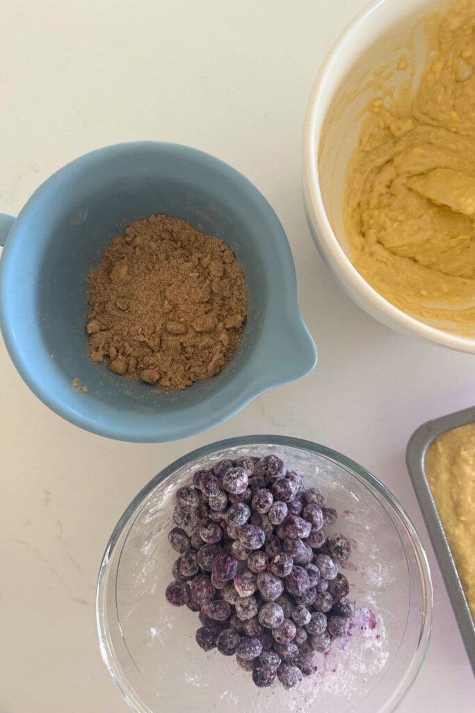A glass bowl of frozen blueberries tossed in flour alongside a blue batter jug containing a brown sugar cinnamon streusel mixture.
