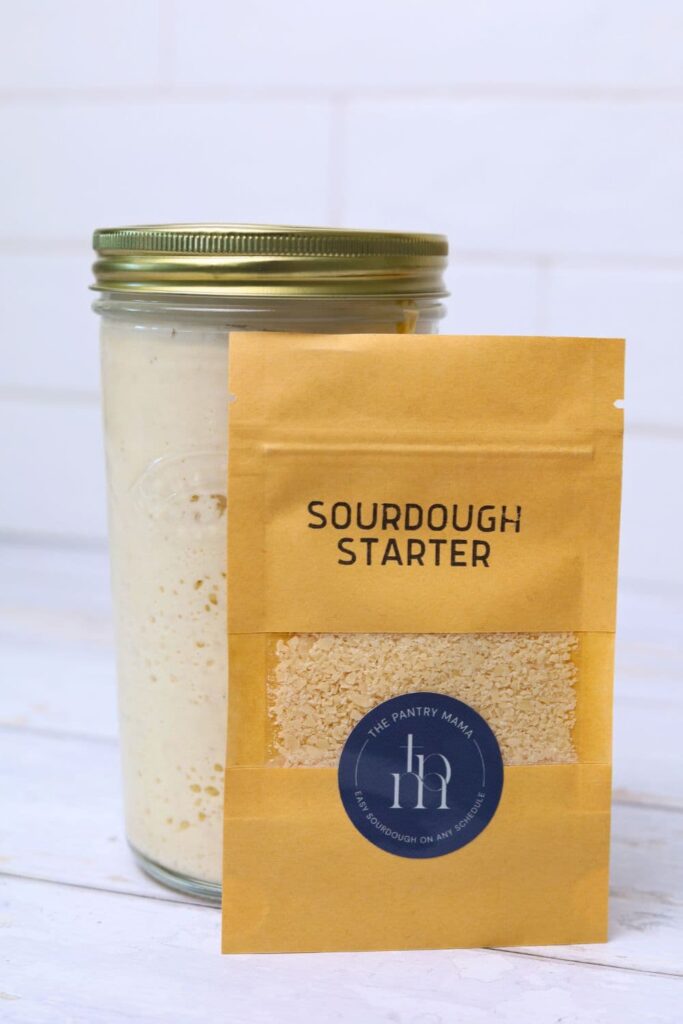 A jar of sourdough starter with a sachet of dried sourdough starter standing in front it. 