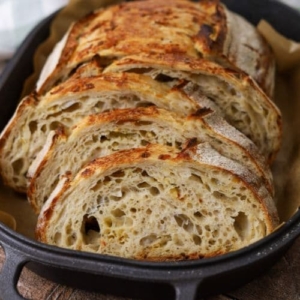 A loaf of Italian herb and cheese sourdough bread that has been sliced and displayed in a cast iron bread pan. This is the recipe feature image.