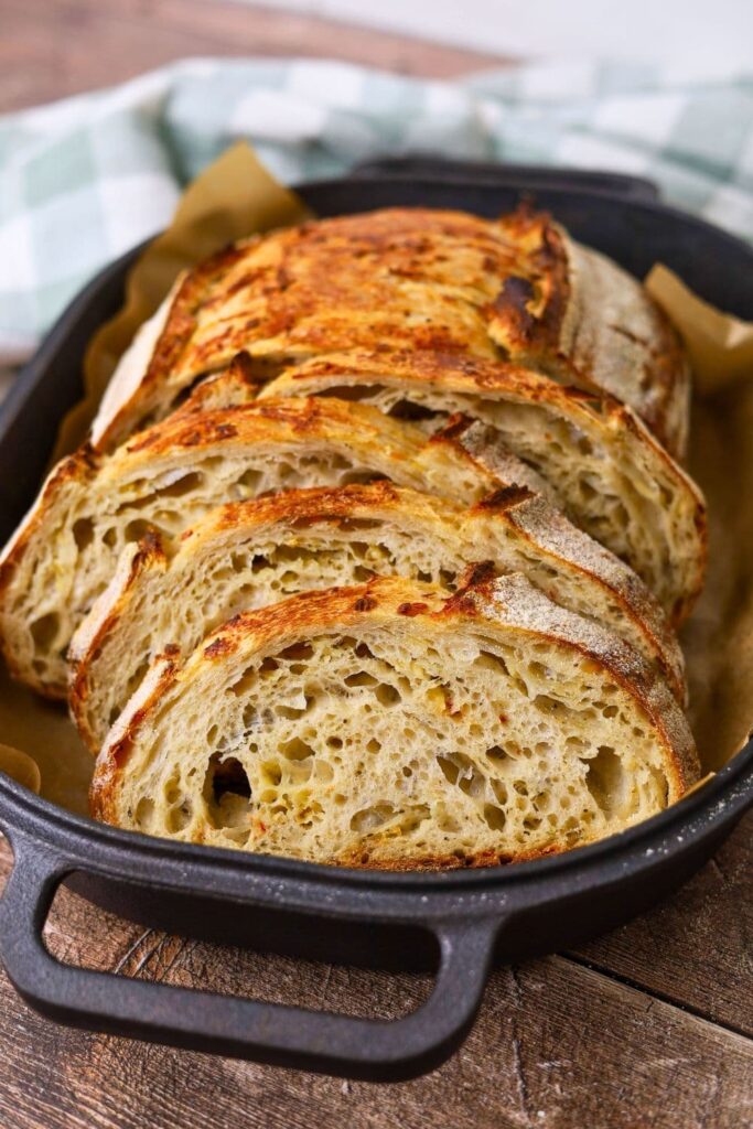 A loaf of sourdough with Italian herbs and cheese that has been sliced and laid out in a black cast iron bread pan.