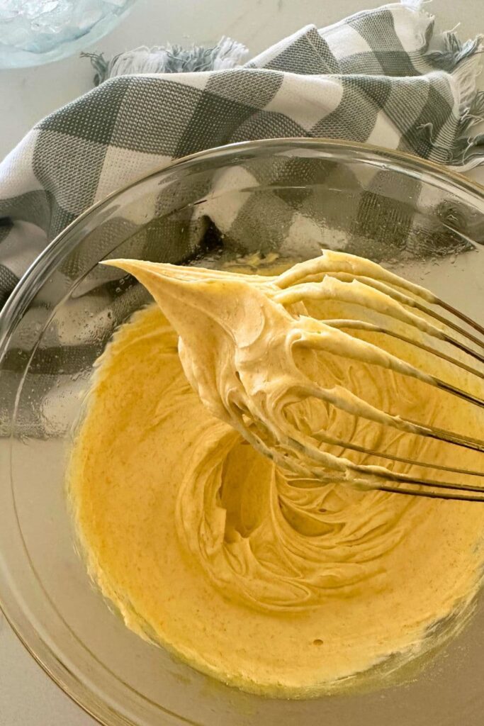 A bowl of whipped brown butter that is thick and creamy. You can see that it is sticking to the whisk. There is a green checked dish towel in the background of the photo.