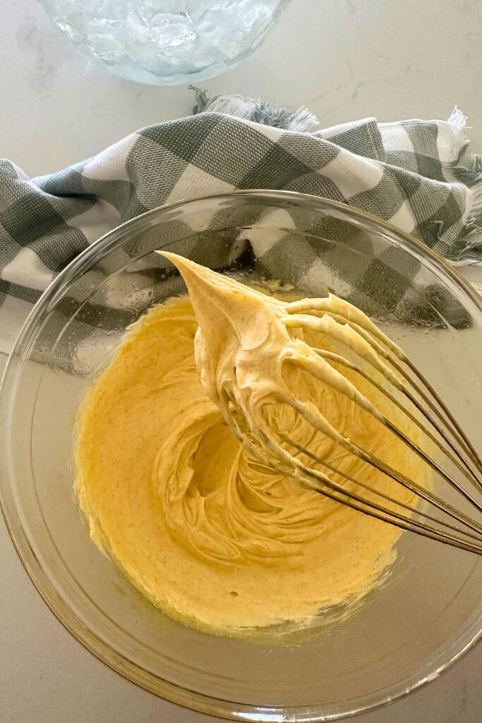 A bowl of whipped brown butter that is thick and creamy. You can see that it is sticking to the whisk. There is a green checked dish towel in the background of the photo.