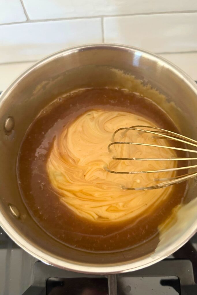 A saucepan containing a caramelized mixture of butter and sugar that has had a can of condensed milk poured on top. There is a whisk that is starting to bring the mixture together.