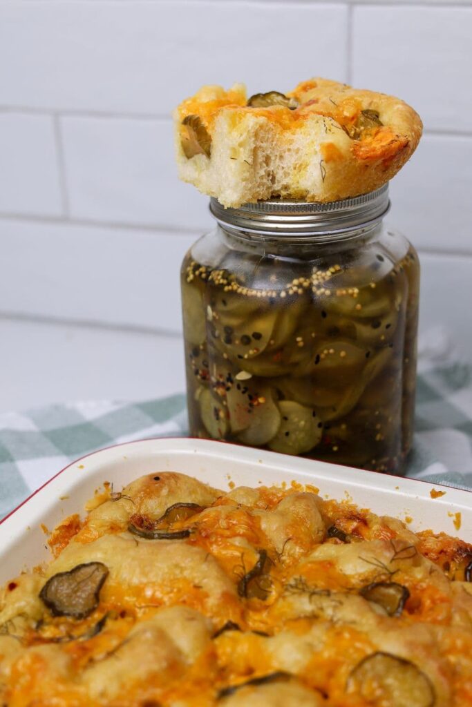A jar of dill pickles with a slice of sourdough dill pickle focaccia sitting on top of the jar. There is an enamel tray of focaccia bread sitting in front of the jar.