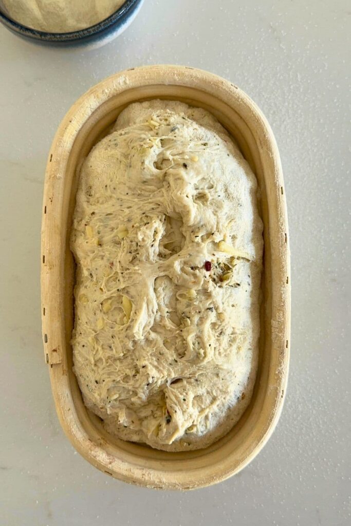 An Italian herb and cheese sourdough loaf that has been placed into a banneton. The top of the dough has started to come apart and needs to be stitched together.