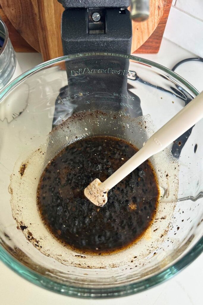 A glass KitchenAid bowl filled with the liquid ingredients for sourdough brown bread that have been stirred together to make a dark molasses slurry. There is a white spatula in the mixture.