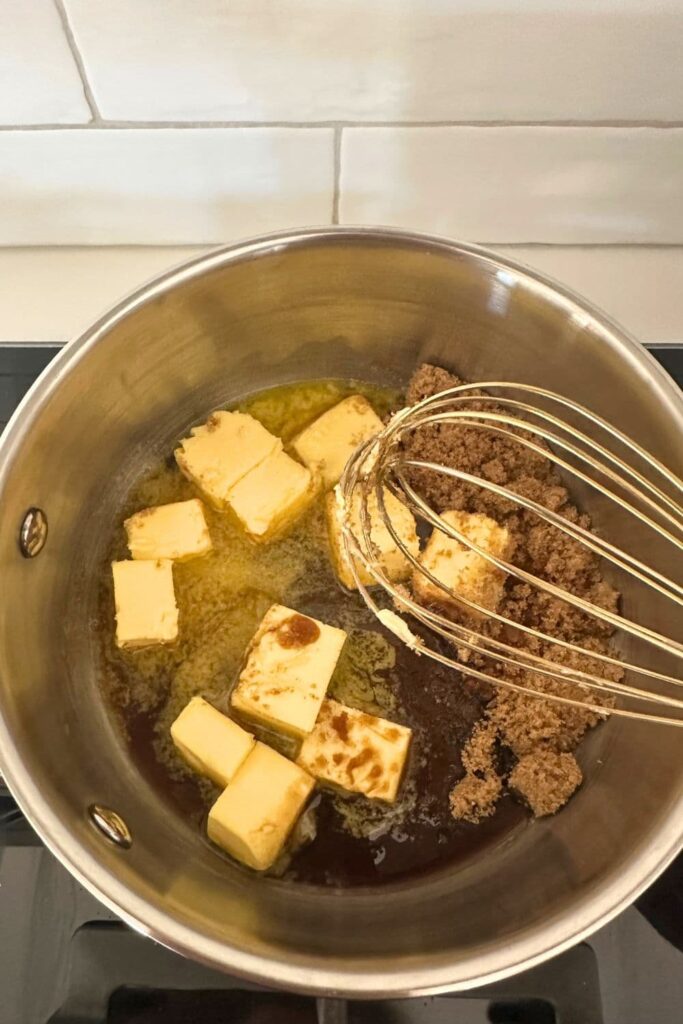 A saucepan containing cubes of butter that are starting to melt with brown sugar. There is a whisk resting on the side of the saucepan.