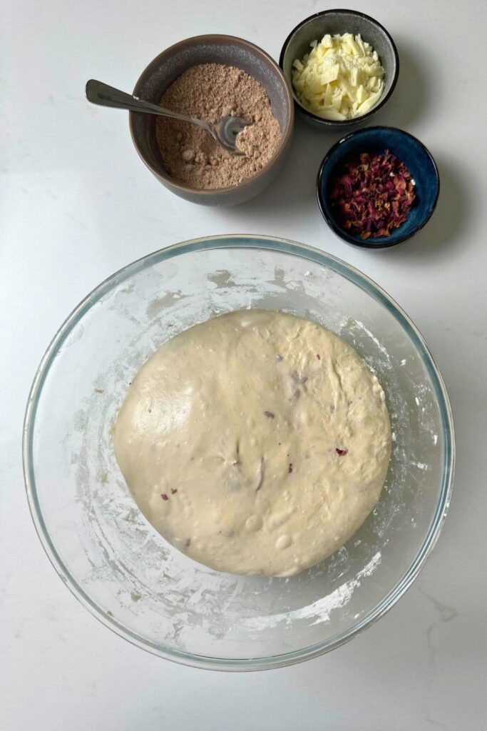 A bowl of rose petal sourdough that has finished bulk fermentation and is ready for shaping. The inclusions that will be added during shaping are in small bowls at the top  of the photo (cinnamon, brown sugar, white chocolate and rose petals).