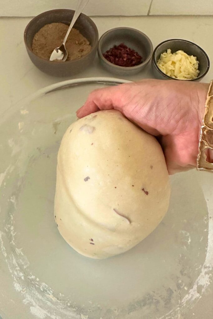 A smooth ball of sourdough infused with rose petals being held by a hand over a bowl. There are three bowls in the background containing cinnamon, white chocolate and rose petals.