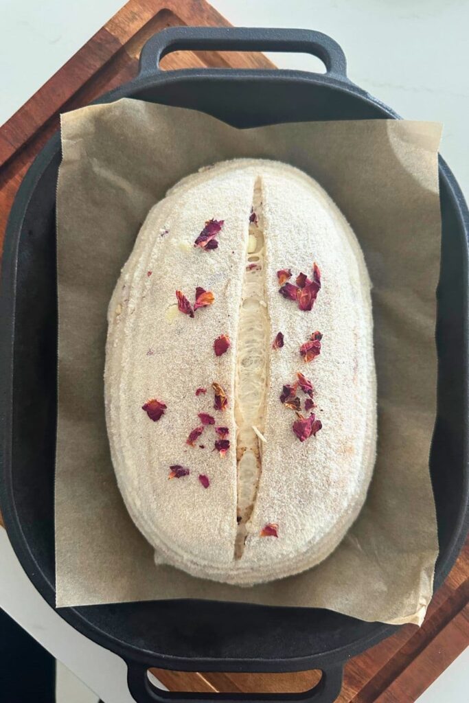 An unbaked loaf of sourdough bread that has been sprinkled with dried rose petals and placed into a hot cast iron bread pan with the lid off.