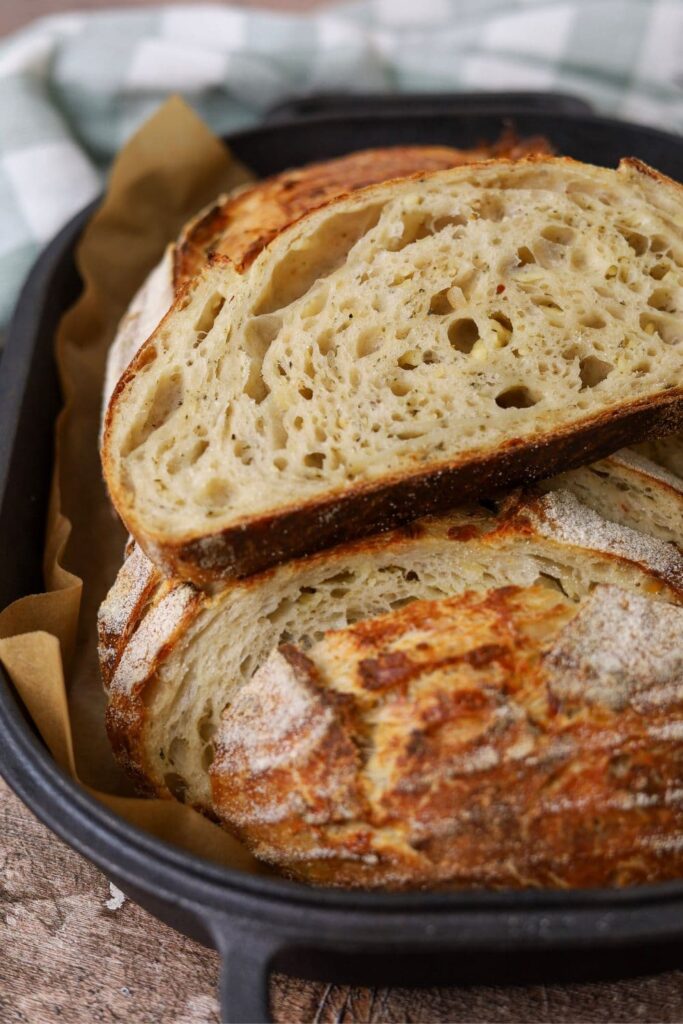 A loaf of sourdough Italian herb and cheese that has been sliced and then placed back into the black cast iron bread pan so you can see not only the inside crumb but also the gorgeous crunchy crust.